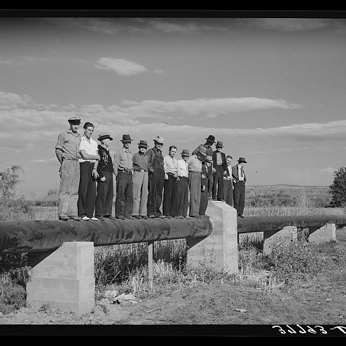 Old, black and white photo of 13 farmers standing on irrigation pipe